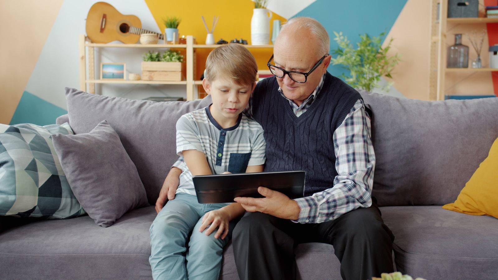 Un grand-père et son petit-fils assis sur un canapé regardent ensemble une tablette.